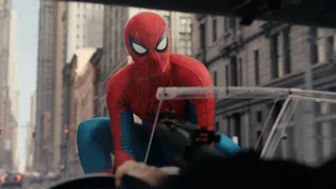 Marvel/ Sony Pictures Entertainment Spiderman on the bonnet of a car, with a number of tall stone buildings behind him.