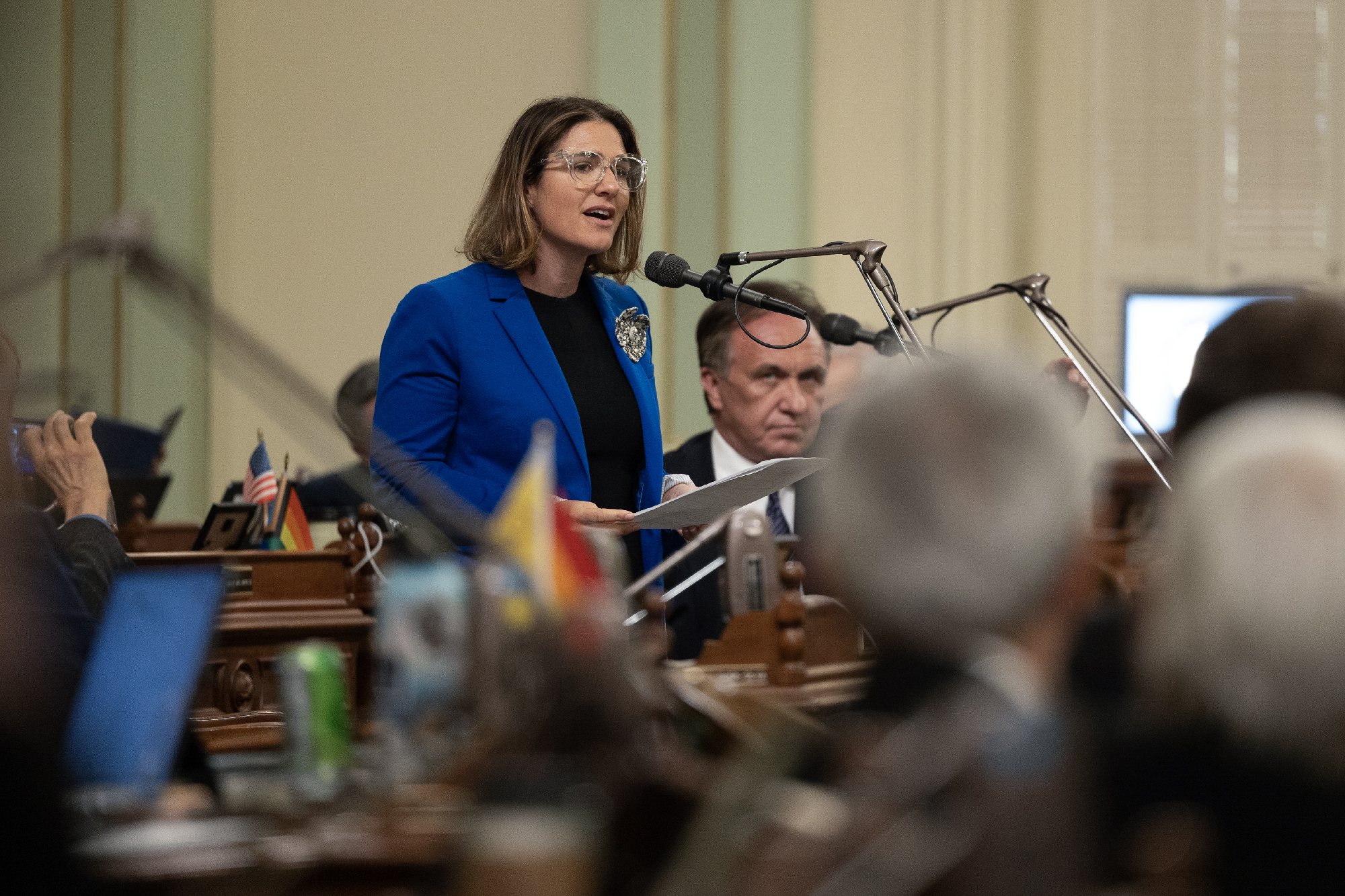 A white woman wearing glasses and blue jacket stands in front of a podium with a microphone around other people.