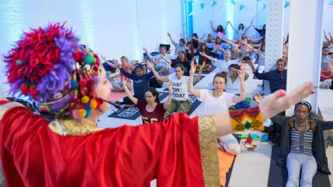 Mind, Body and Spirit Festival A woman is wearing a red and purple coloured hat along with a red shawl. She has her arms pointed outwards to the sky. People are sitting on the floor on yoga mats, copying her arm actions.