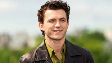 Getty Images Tom Holland smiling in an olive green shirt and brown leather jacket standing in front of some green foliage in the distance and a blue sky in a photoshoot for the film The Odyssey in London