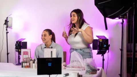 Daisy Kelly Two young women surrounded by studio lights and tripods selling eyelash serums on a live stream