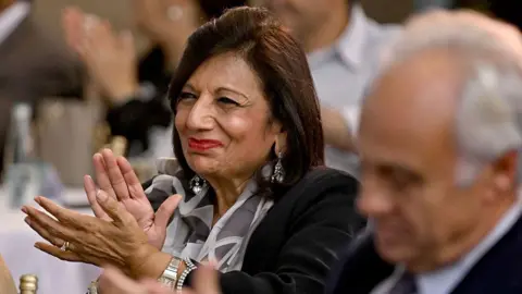 AFP via Getty Images Kiran Mazumdar-Shaw, Indian billionaire entrepreneur and founder of Biocon Limited pictured with a smile, clapping her hands, sitting next to an older gentleman.