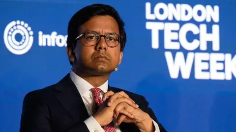 Getty Images Kanishka Narayan looks pensive with his hands folded together and a microphone positioned by his chin as he sits on stage at London Tech Week. The conference's logo is displayed on a backdrop behind him.