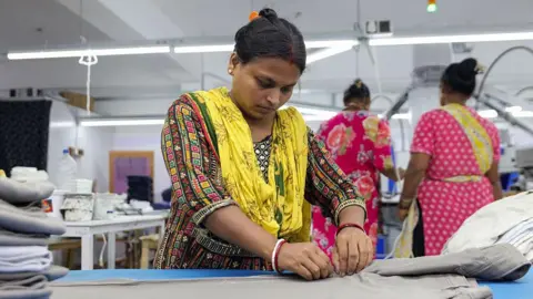 NurPhoto via Getty Images A woman wearing a multi-coloured Indian kurta with a yellow stole, finishes men's trousers after production in Kolkata city, India,.