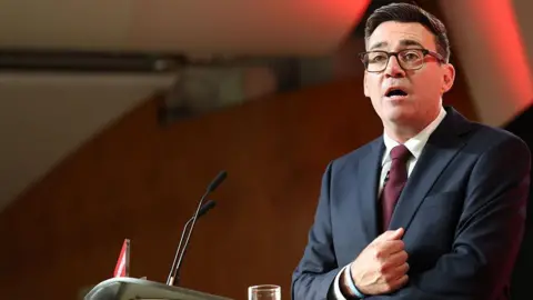 Getty Images Andy Burnham stood at a lectern delivering his first speech as the leader of the Labour Party. His right hand is clenched in a fist and pulled towards his chest as he speaks. He is wearing glassed, a black suit, white shirt, and burgundy tie.