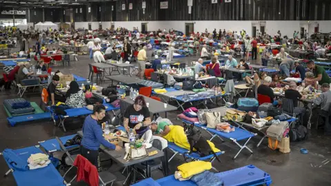 Lorena Sopena Lopez/Anadolu via Getty Images People sit at makeshift table and chairs in a convention centre with supplies on a table