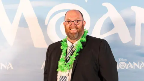 James Gillham/Disney A man wearing a suit and glasses and a green flower necklace. He is smiling while standing in front of a Moana backdrop.