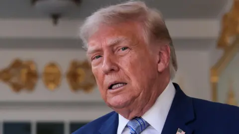 Reuters US President Donald Trump speaking in front of a White House entrance, wearing a blue suit and tie.