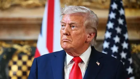 Getty Images A photo of Donald Trump from the shoulders up. He is wearing a navy suit with a white shirt and red tie, with a US flag pin on his lapel. He is looking to the left.
