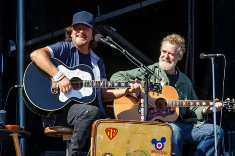 Getty Images Two men are seated on a stage, both with acoustic guitars and in front of microphones. The man on the left is earing a blue shirt and blue baseball cap, he is smiling. His guitar is also a dark blue. The man on the left is wearing a green shirt and blue jeans. He is also smiling. It is daytime.