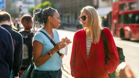Getty Images Two women standing in the street talking. One is wearing a blue t shirt and the other is wearing a red coat