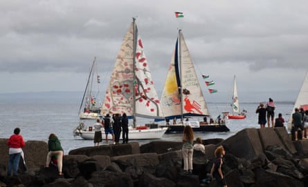 People stand on rocks watching a group of small sailing boats, some with brightly painted and decorated sails and flying Palestinian flags.