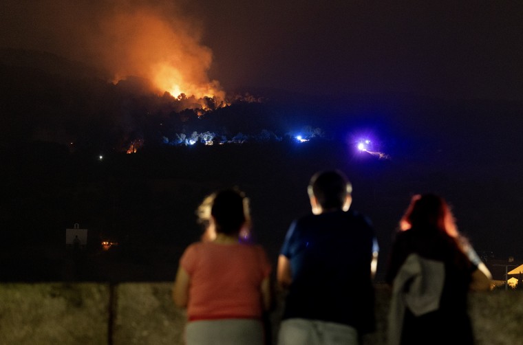 Three people are silhouetted as they face toward a distant wildfire at night.