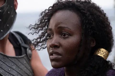 Universal Pictures Lupita Nyong'o looks to the left of frame in a concerned manner, in a close head and shoulders shot as she stars as Helen of Troy in Christopher Nolan's The Odyssey.