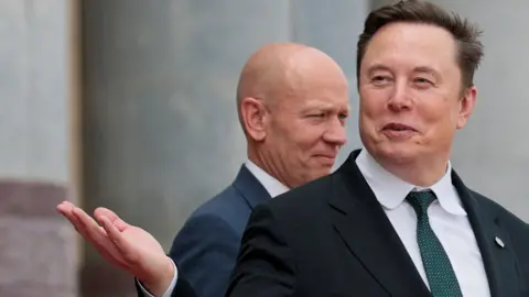 Reuters Elon Musk leaving a building in China, smirking and gesturing with his hand while wearing a black suit jacket, white collared shirt and dark green tie.