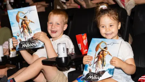 James Gillham/Disney Two children holding up Moana posters while sitting in cinema seats