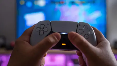 Getty Images A white PlayStation 5 controller being held up by someone's hands against a blurred TV screen behind it