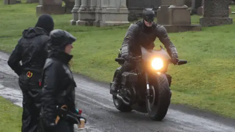 PA Media Filming on a scene in The Batman - a man dressed in a Batman costume is riding a motorbike in a graveyard, watched by crew members