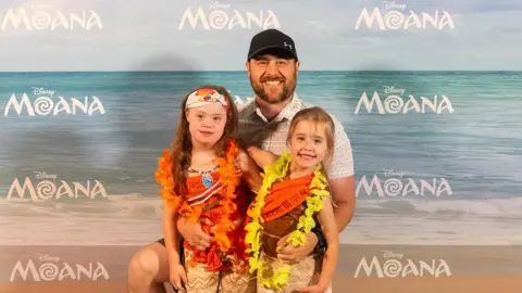 James Gillham/Disney Two children with Moana outfits and colourful floral necklaces on standing in front of a man wearing a shirt and a hat. They are all in front of a Moana backdrop.