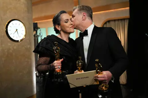 Getty Images On the right -- Christopher Nolan holding two Oscars and Best Picture envelope, in black dinner jacket kissing his wife on the cheek
On the left - Emma Thomas in black dress holding Oscar