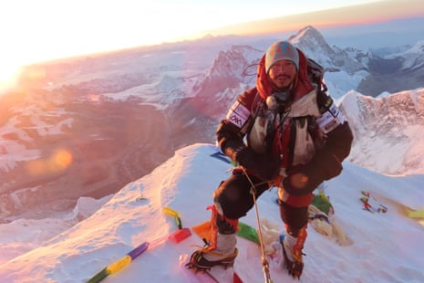 Nirmal Purja in an orange jacket stands on a snowy summit at sunrise with mountain peaks behind