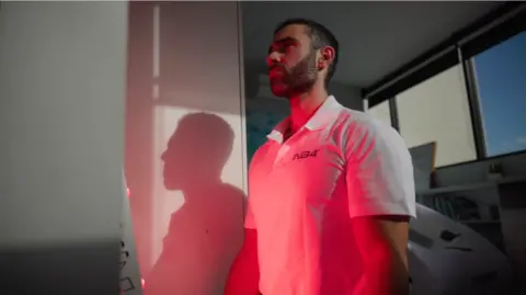 Lucas stands indoors with a red light projecting onto his body. He has short dark hair with a dark beard. He is wearing a white polo-neck t-shirt.