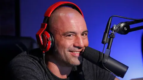 Getty Images Joe Rogan, wearing orange headphones over his ears, smiles into a microphone during a taping of Joe Rogan Questions Everything