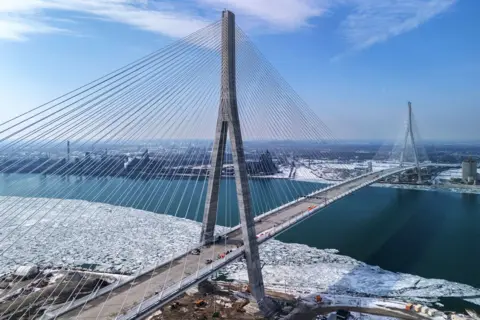 Dax Melmer / Reuters The Gordie Howe International Bridge, a cable-stayed bridge featuring six traffic lanes, is seen in a drone image. The ground on either side is covered in snow, with unfrozen water flowing beneath.