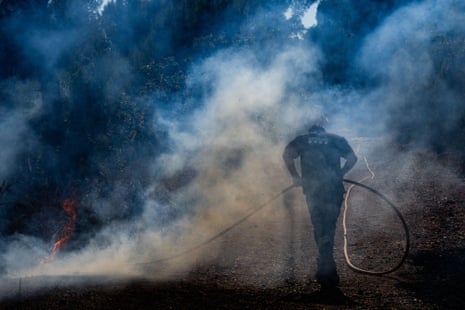 A firefighter holding a hose walks through thick smoke on a road near a burning fire