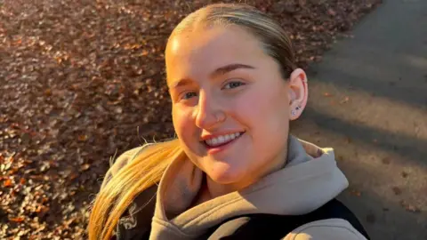 Sophia Picture of a young woman smiling for a selfie in the woods. She has blonde hair slicked back in a ponytail and is wearing a stone coloured hoodie with a black body warmer.