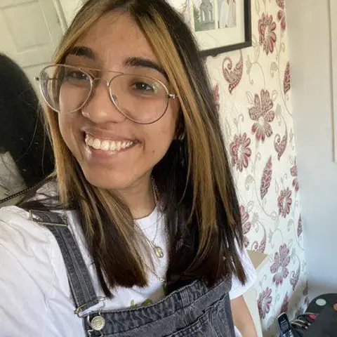 Jazmin Kaur A selfie shot of Jazmin Kaur smiling on camera. She's wearing dungarees and a white t-shirt and has long brown hair and clear glasses.