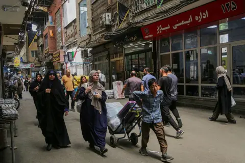 Getty Images Wide shot showing a busy street with shops in Tehran. There are several men and women walking through the street At the front of the image is a young boy wheeling a trolley behind him.