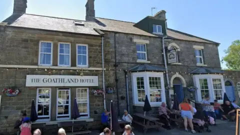 Google The exterior of The Goathland Hotel, a stone-built two-storey structure built from grey stone, with a slate roof and several chimney stacks.
Above the entrance on the left-hand side is a large sign reading THE GOATHLAND HOTEL. The front of the building features large sash-style windows, stone detailing around the openings, and hanging flower baskets.
Multiple visitors are seated or standing in front of the building, with some gathered around the outdoor seating area.