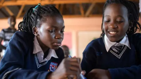 AFP via Getty Images Two 13-year-old girls, who are members of the Ugandan dance group Ghetto Kids, attend school in Kampala on 20 Apri 2023. They are looking down at a book and wearing navy school jumpers and white shirts.