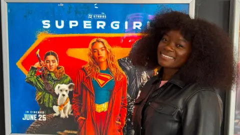 BBC A woman in a black leather jacket is smiling at the camera. She is standing next to a Supergirl film poster