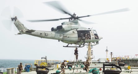 Marines descend from a helicopter which is hovering over the deck of a large ship