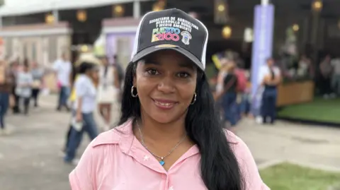 Doralba Bejarano smiles at the camera from a busy public square. She is wearing a black baseball cap with slogans including the name "Puerto Rico", and a pink shirt.