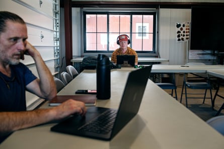 Man in T-shirt sits at laptop at long table, with second man in headphones at laptop at adjoining table.