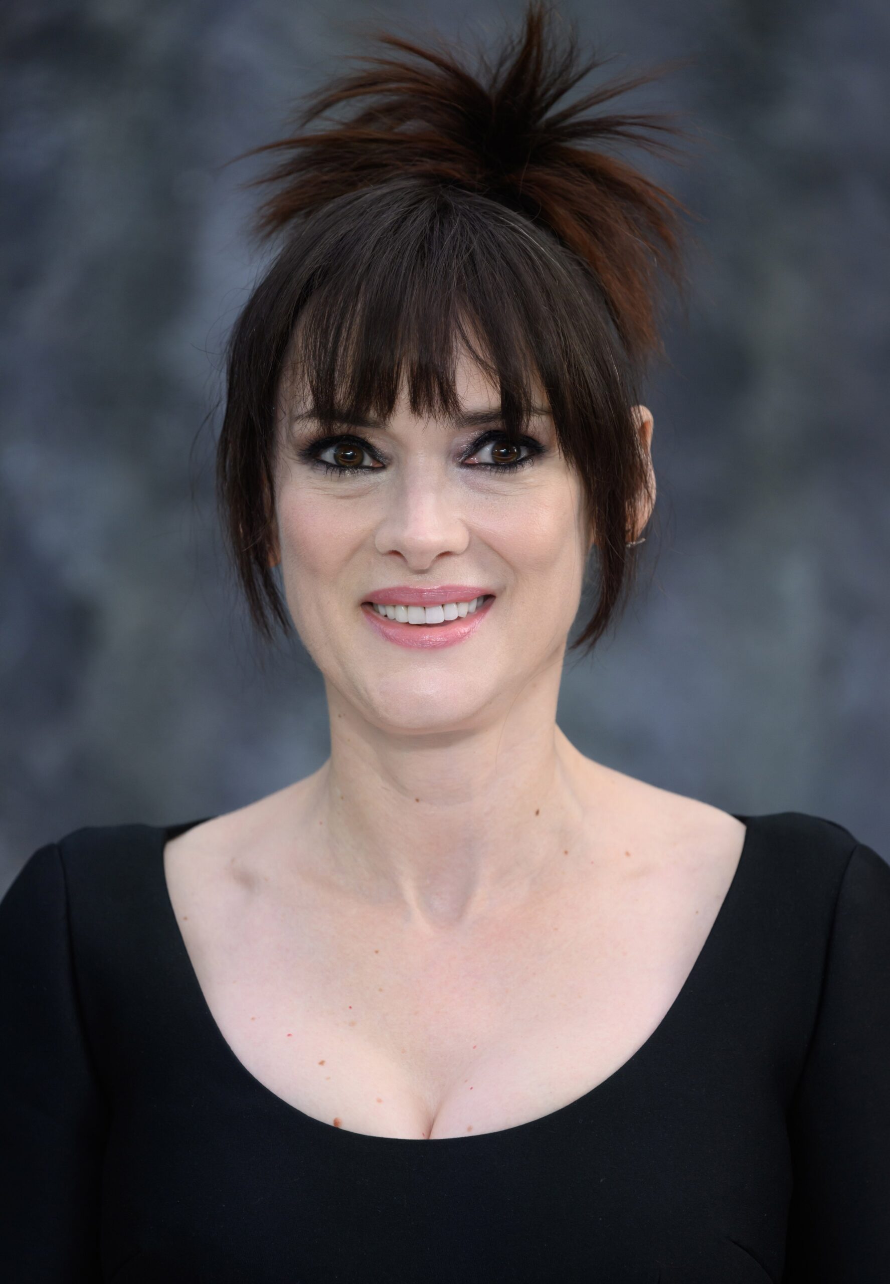 Winona Ryder with an updo hairstyle and bangs, wearing a simple, elegant outfit, on a background