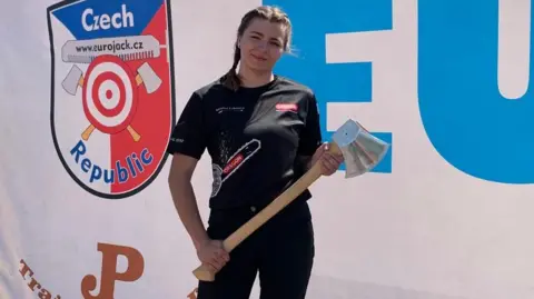 Karolina Urbanova Karolina Urbanova squints and smiles into the sun while holding an axe and wearing her hair in two braids