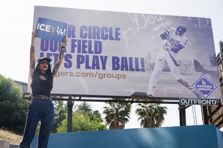 a person holds a sign that reads ‘ice out of LA’ in front of a billboard for the Los Angeles Dodgers
