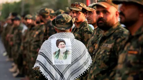 AFP A member of Iraq's Popular Mobilisation Forces (PMF) wears a keffiyeh scarf with a portrait of Iran's late Supreme Leader Ali Khamenei stuck to it, at the shrine of Imam Hussein on the eve of his funeral ceremonies in the holy Iraqi city of Karbala (7 July 2026)