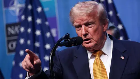 NurPhoto via Getty Images US President Donald Trump wearing a yellow tie, white shirt, blue suit and American flag pin. He stands at a microphone and is pointing, his mouth open.
