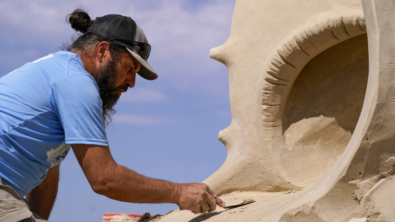 Man in blue shirt and gray baseball cap scrapes sand off sculpture using garden tool