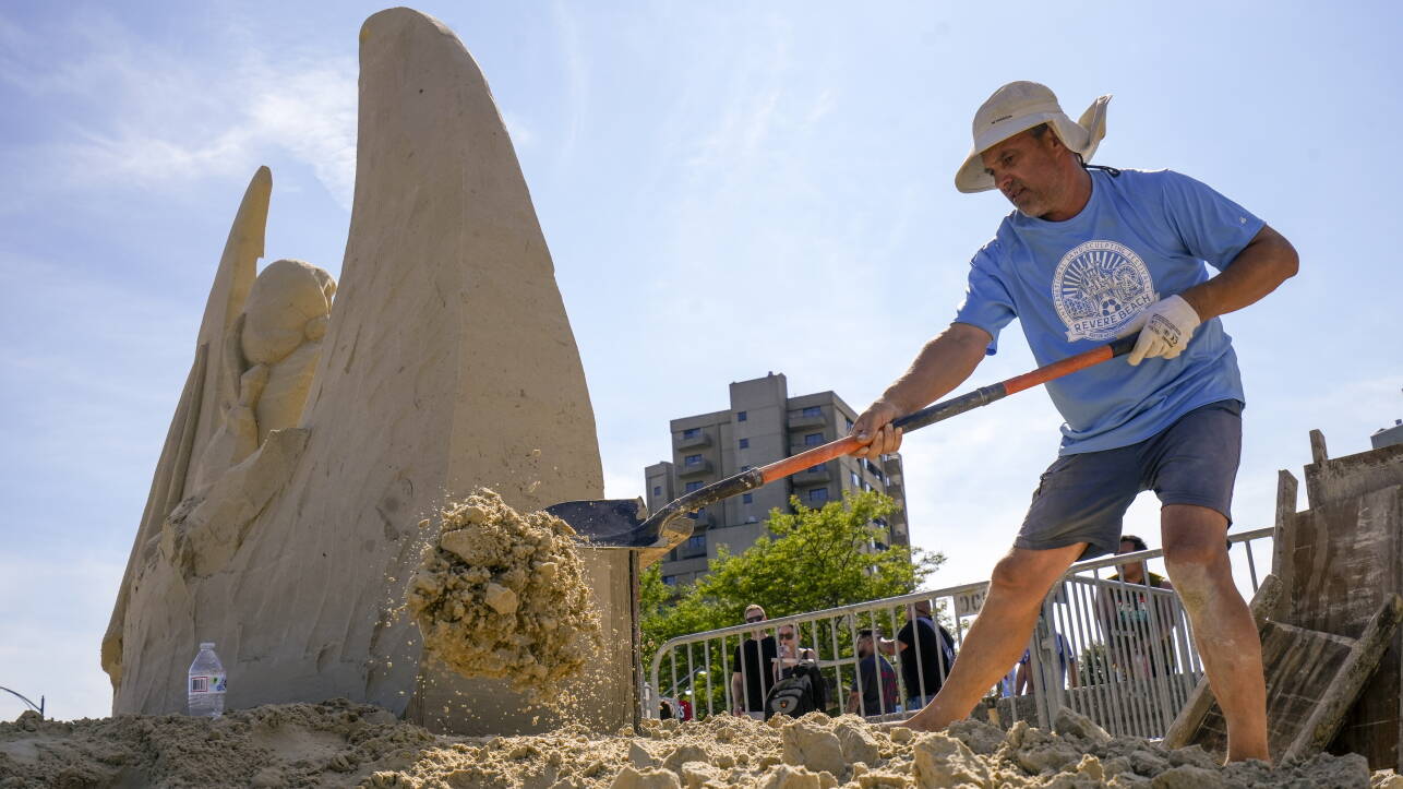 Man in blue tee shirt and navy shorts shovels pile of sand with metal shovel