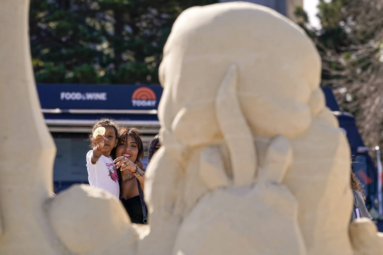 Girl in white shirt held by woman in black shirt points at sand sculpture in foreground