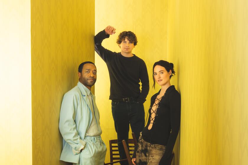 Three people stand in a yellow hallway with a jumble of chairs.