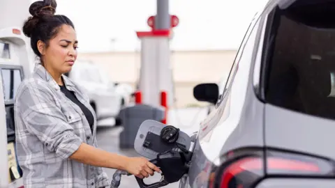 Getty Images Woman wearing a grey shirt fills up her car at a gas station