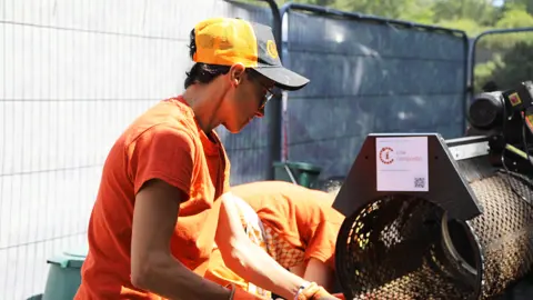 Heledd Wyn ffin with an orange and black baseball style cap on and an orange Criw Compostio t-shirt. She is wearing glasses and appears to be holding something in her hands out of the picture, possibly a shovel, and putting something into the circular opening of a composting or shredding machine to her side, which has a Criw Compostio label on the top of it. She is photographed from the side and looking down at what she is doing. Behind her to the right Steph, also wearing orange, is bent over and her head is out of sight behind the machine. There is movable fencing behind them.