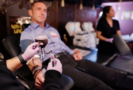 a person with gloved hands holds a vial hooked up to a bag with blood flowing into it from a man seated in a chair